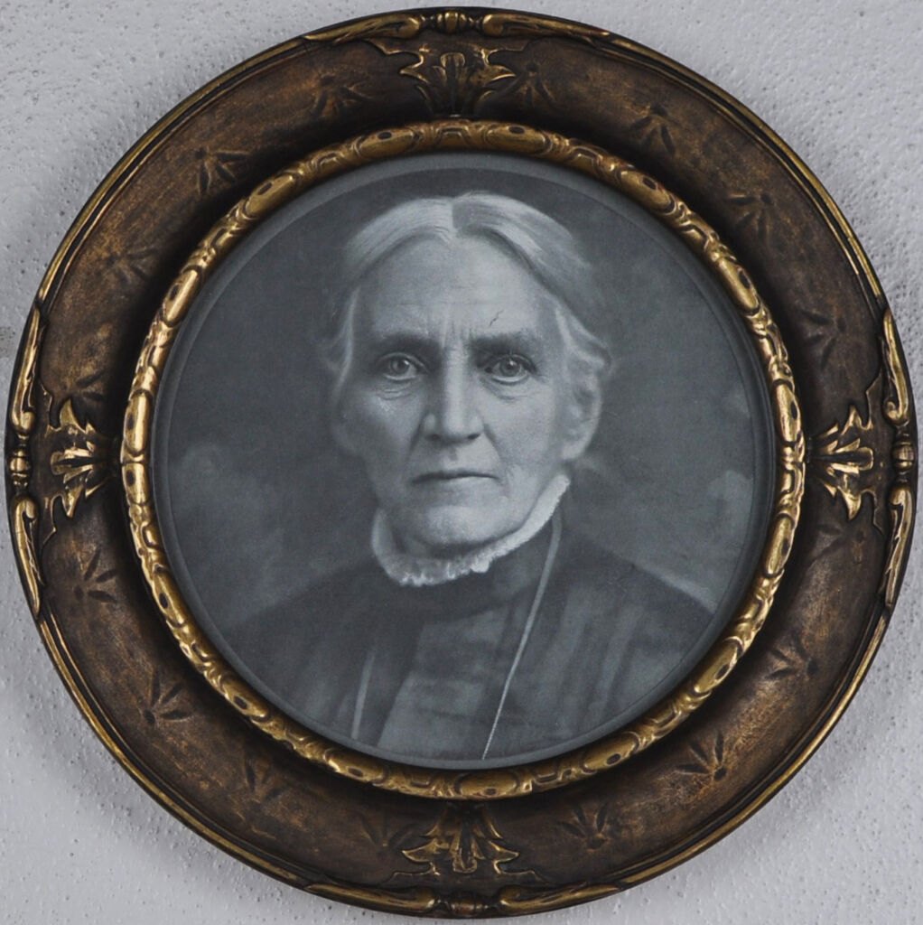 Black and white photograph in a round wood frame. Photo depicts the head and shoulders of an elderly white woman with pulled back hair and a high-necked black dress.